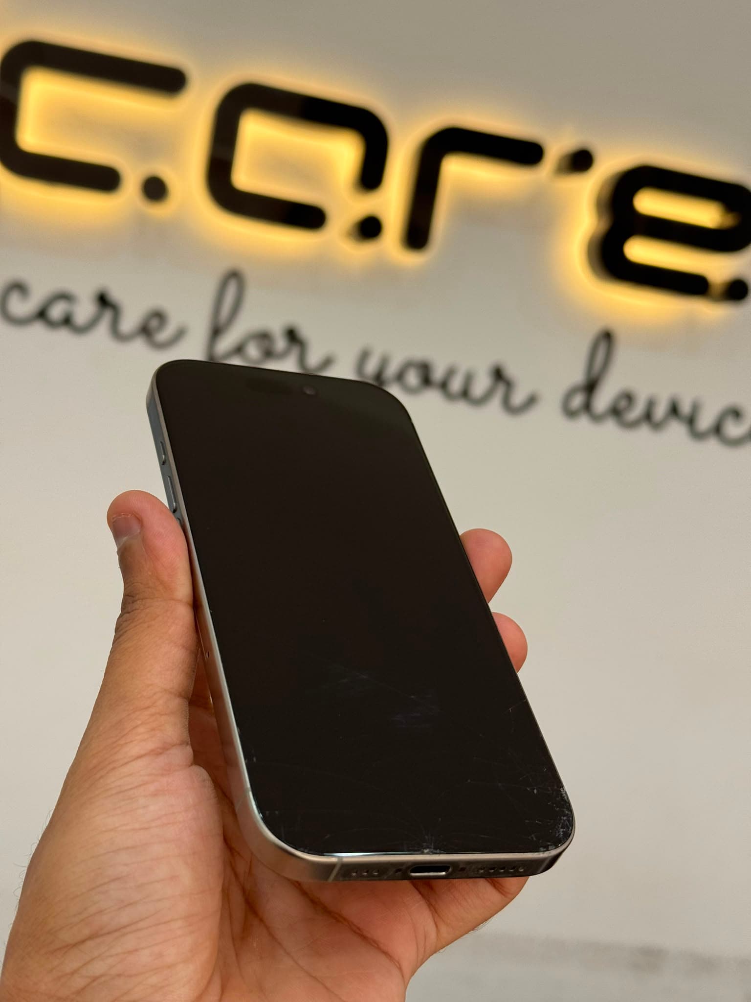 iPhone 16 Pro before screen repair, held in front of the ICARE sign at the Al Gharafa workshop