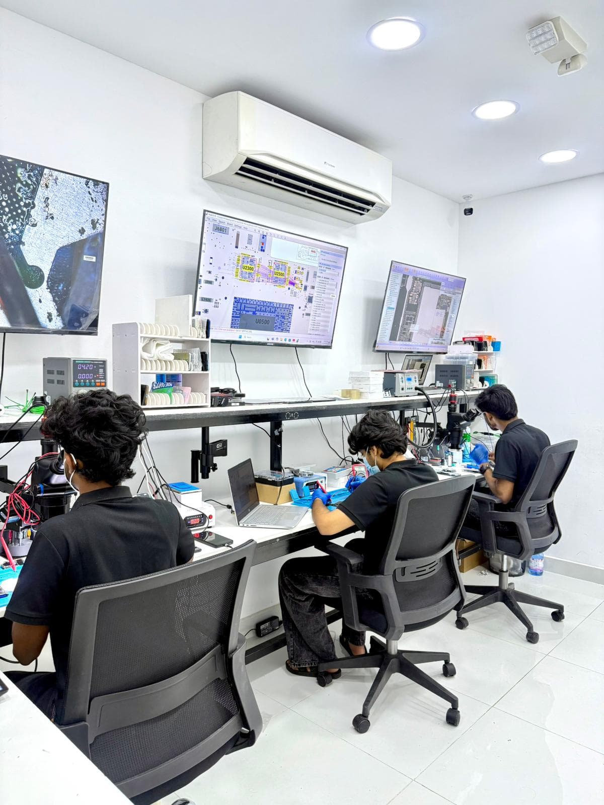 ICARE technicians repairing devices at board-level diagnostic workbenches, with microscopes and repair-schematic monitors, at the Al Gharafa workshop in Doha